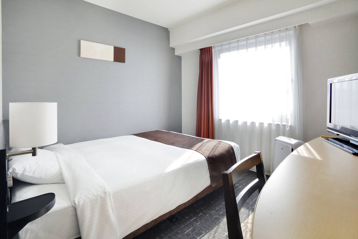 A hotel room with a bed, a desk, a chair, a lamp, a television, and a window with curtains.