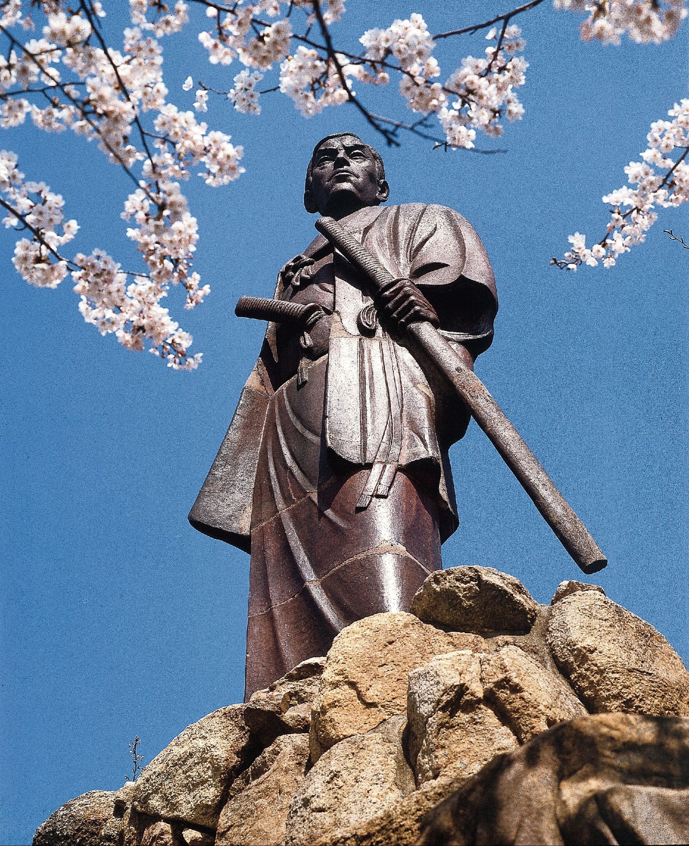 En statue af en figur med en stav, foran en klar blå himmel med kirsebærblomster.