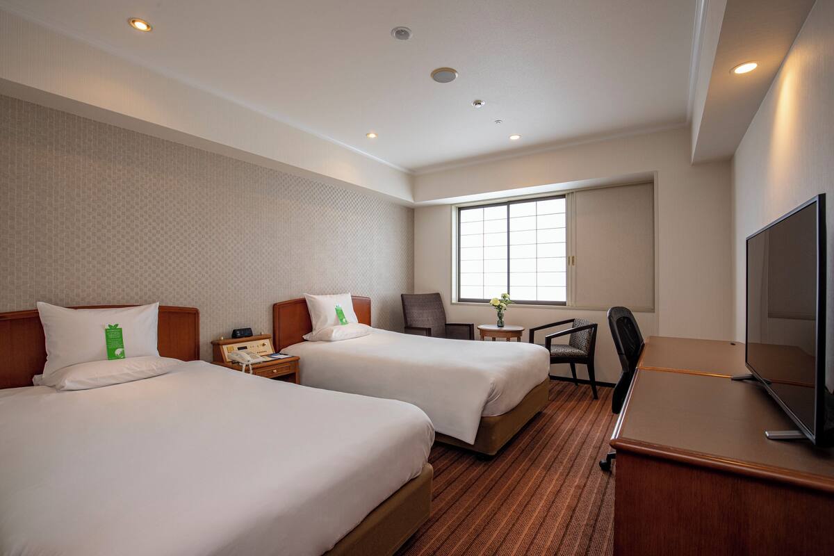 A hotel room with two beds, a desk, a chair, a TV, and a window.