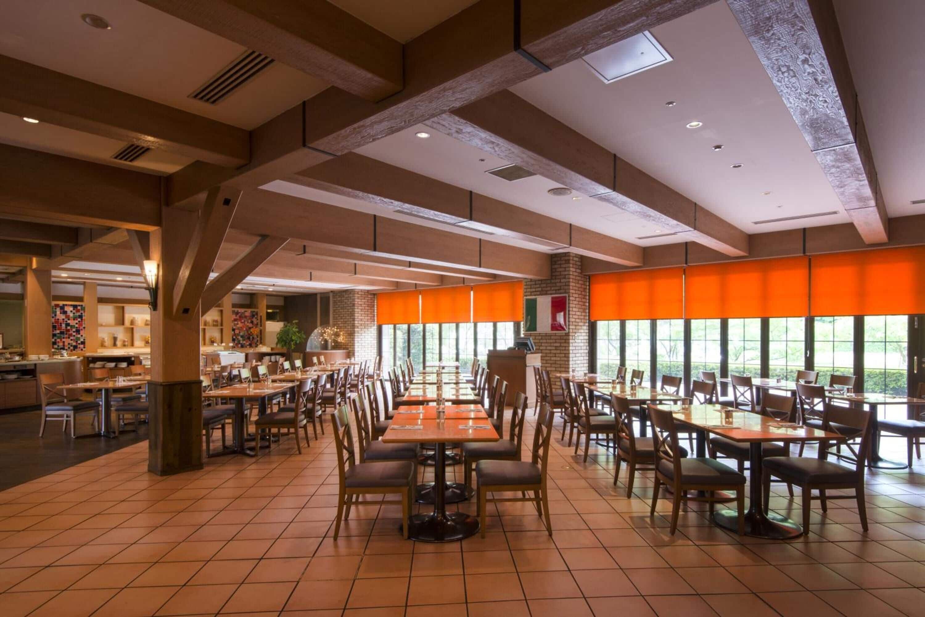 Un'ampia sala da pranzo con diversi tavoli e sedie, grandi finestre con tende arancioni e un moderno design del soffitto.