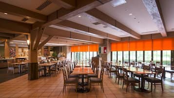 A spacious dining area with multiple tables and chairs, large windows with orange curtains, and a modern ceiling design.