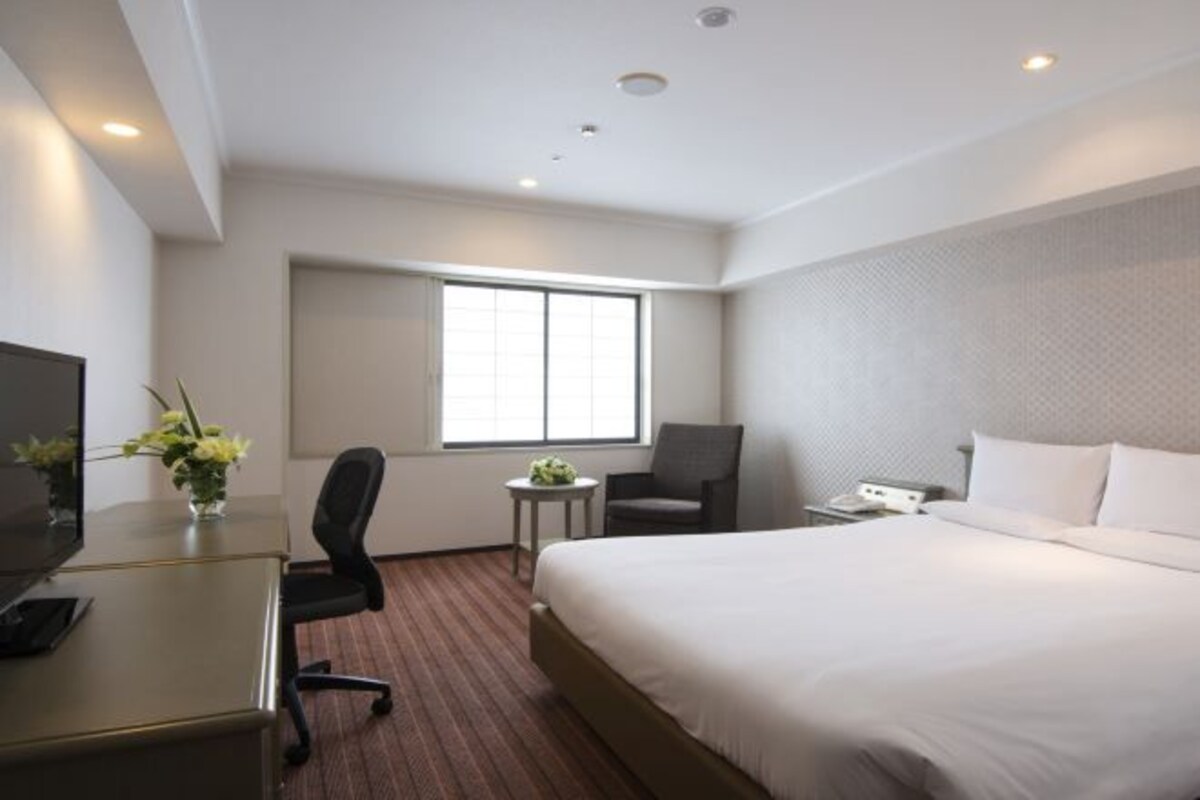Habitación de hotel con cama, escritorio con computadora, silla, una mesita con planta y ventana que deja entrar luz natural.