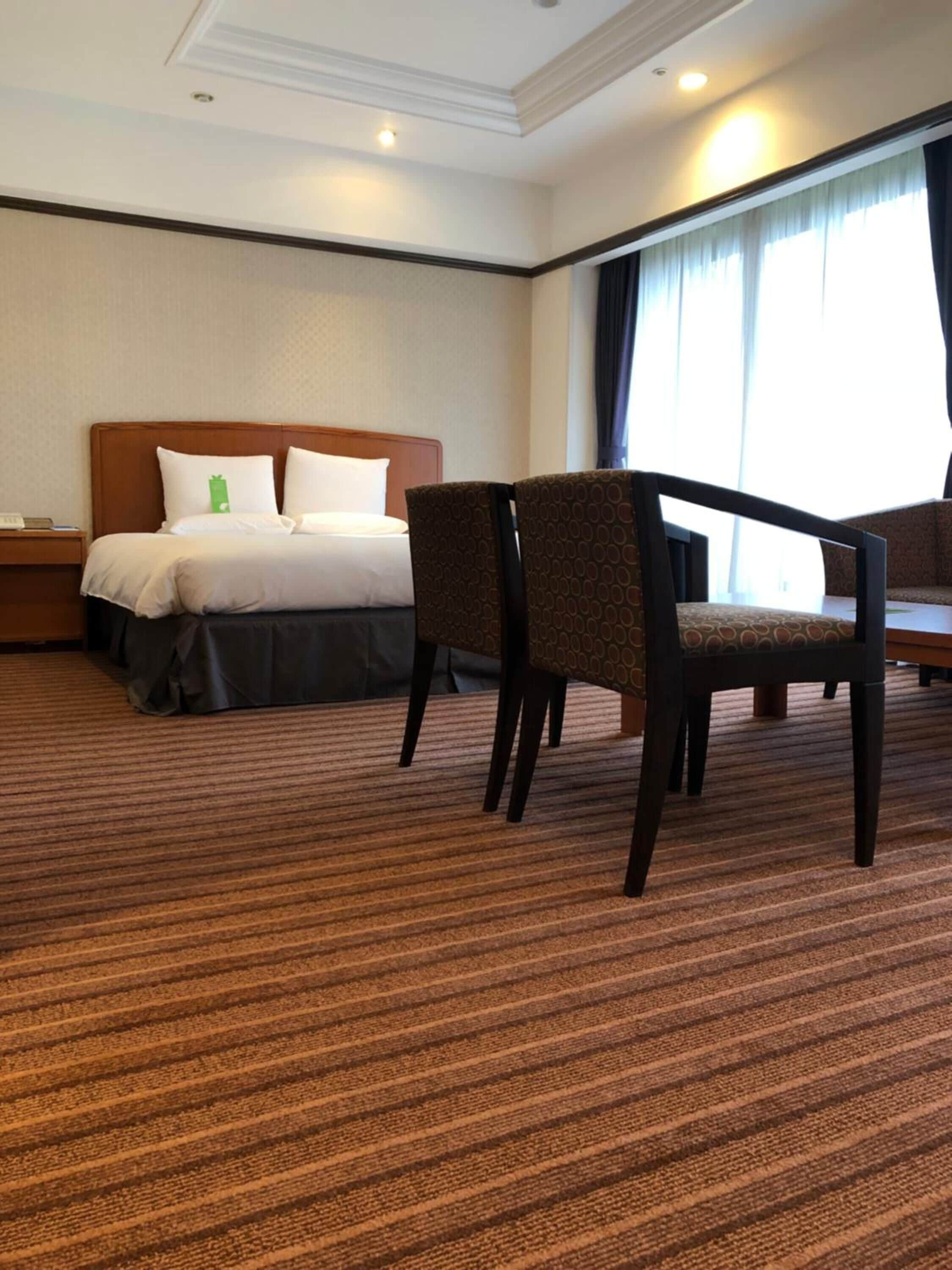 A hotel room with a bed, two chairs, a nightstand, and a large window with curtains.