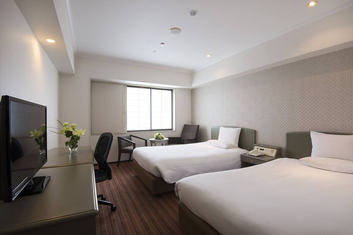 A hotel room with two beds, a desk with a computer, a TV, and a window.