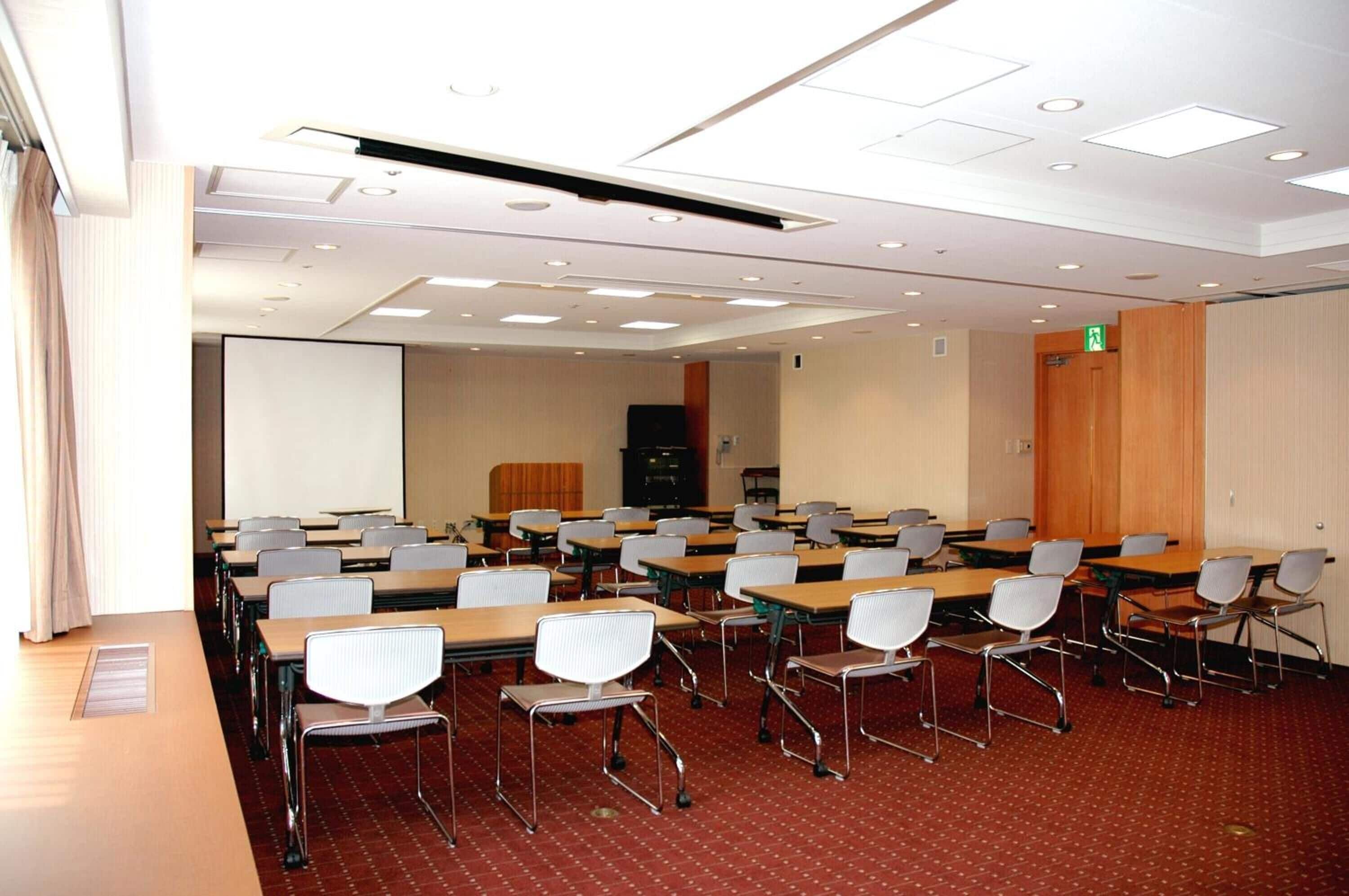 A conference room with rows of tables and chairs, a projector screen, and a wooden panel wall.