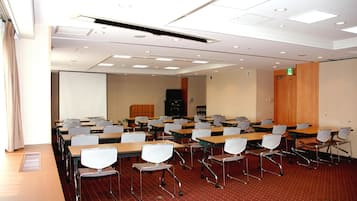 A conference room with rows of tables and chairs, a projector screen, and a wooden panel wall.