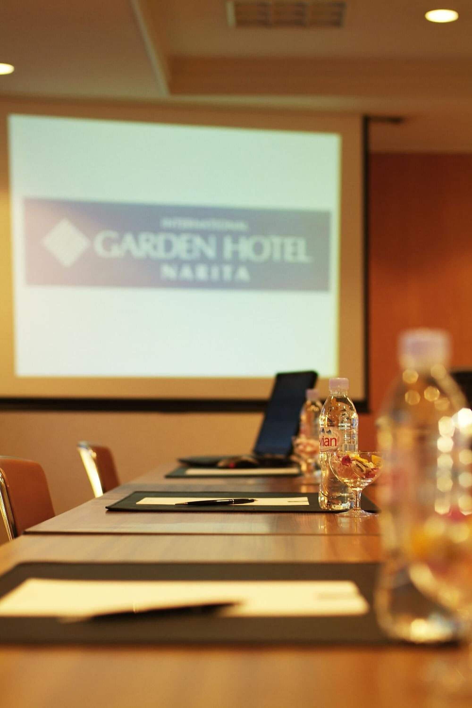 Sala de conferência com uma mesa comprida, garrafas de água e uma tela de projeção exibindo o logotipo do Garden Hotel.