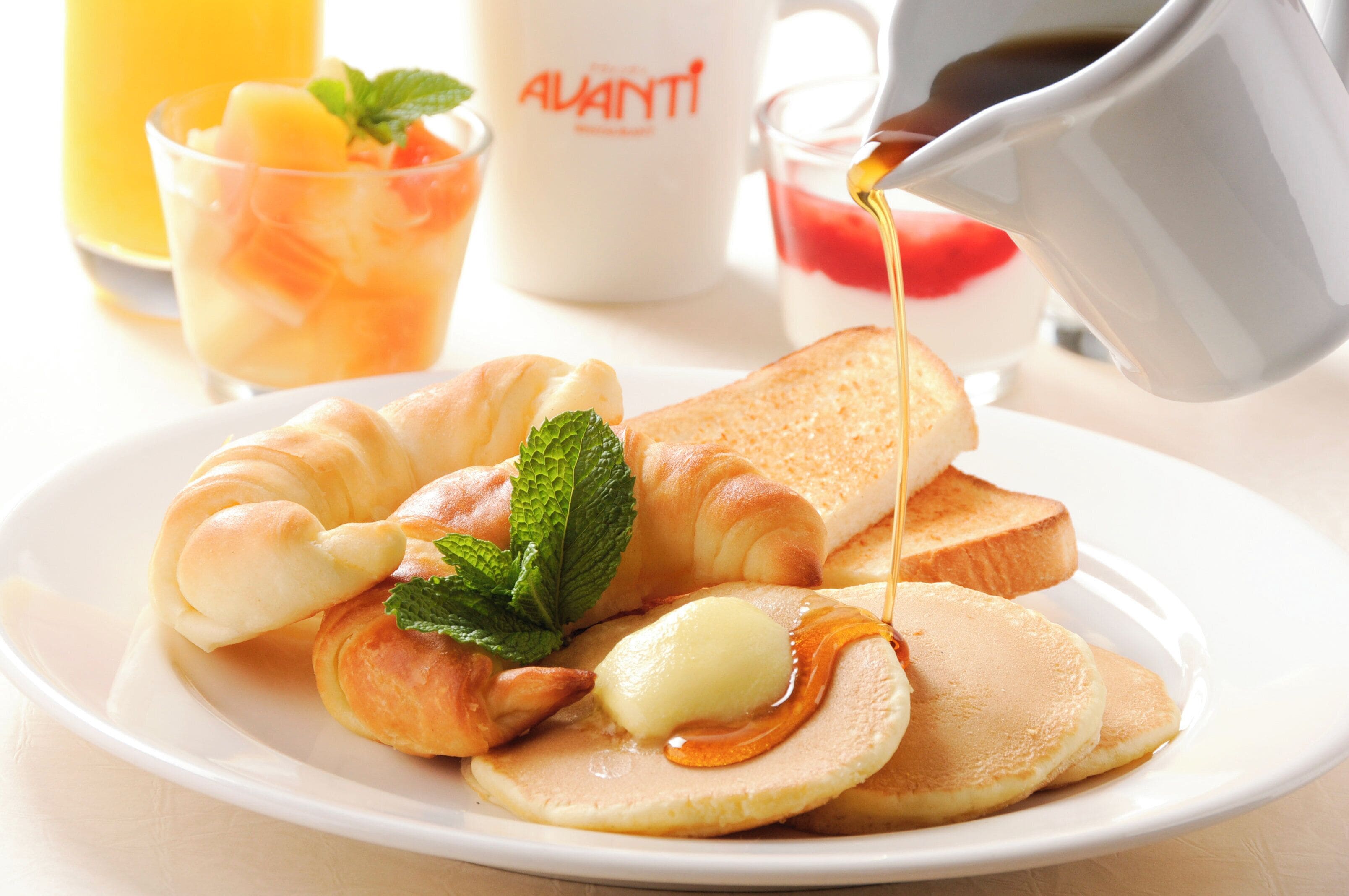 Une assiette de petit-déjeuner comprenant des croissants, des toasts, des crêpes et du sirop, accompagnée d’un verre de jus d’orange et d’une tasse portant l’inscription « ALVANTI ».