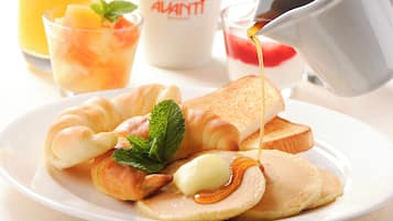 Une assiette de petit-déjeuner comprenant des croissants, des toasts, des crêpes et du sirop, accompagnée d’un verre de jus d’orange et d’une tasse portant l’inscription « ALVANTI ».