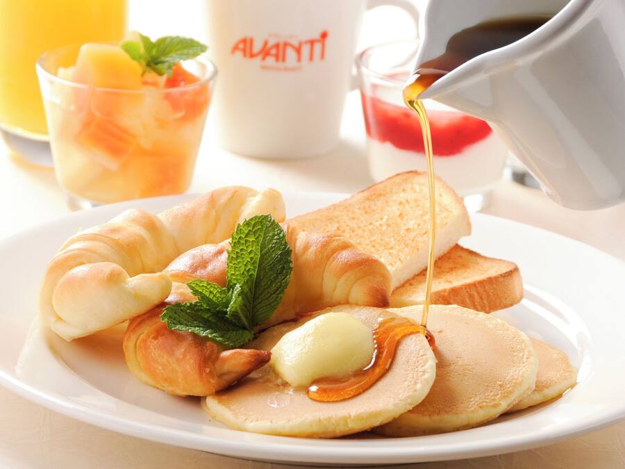 Ein Frühstücksteller mit Croissants, Toast, Pfannkuchen und Sirup, dazu ein Glas Orangensaft und eine Tasse mit dem Schriftzug "ALVANTI".