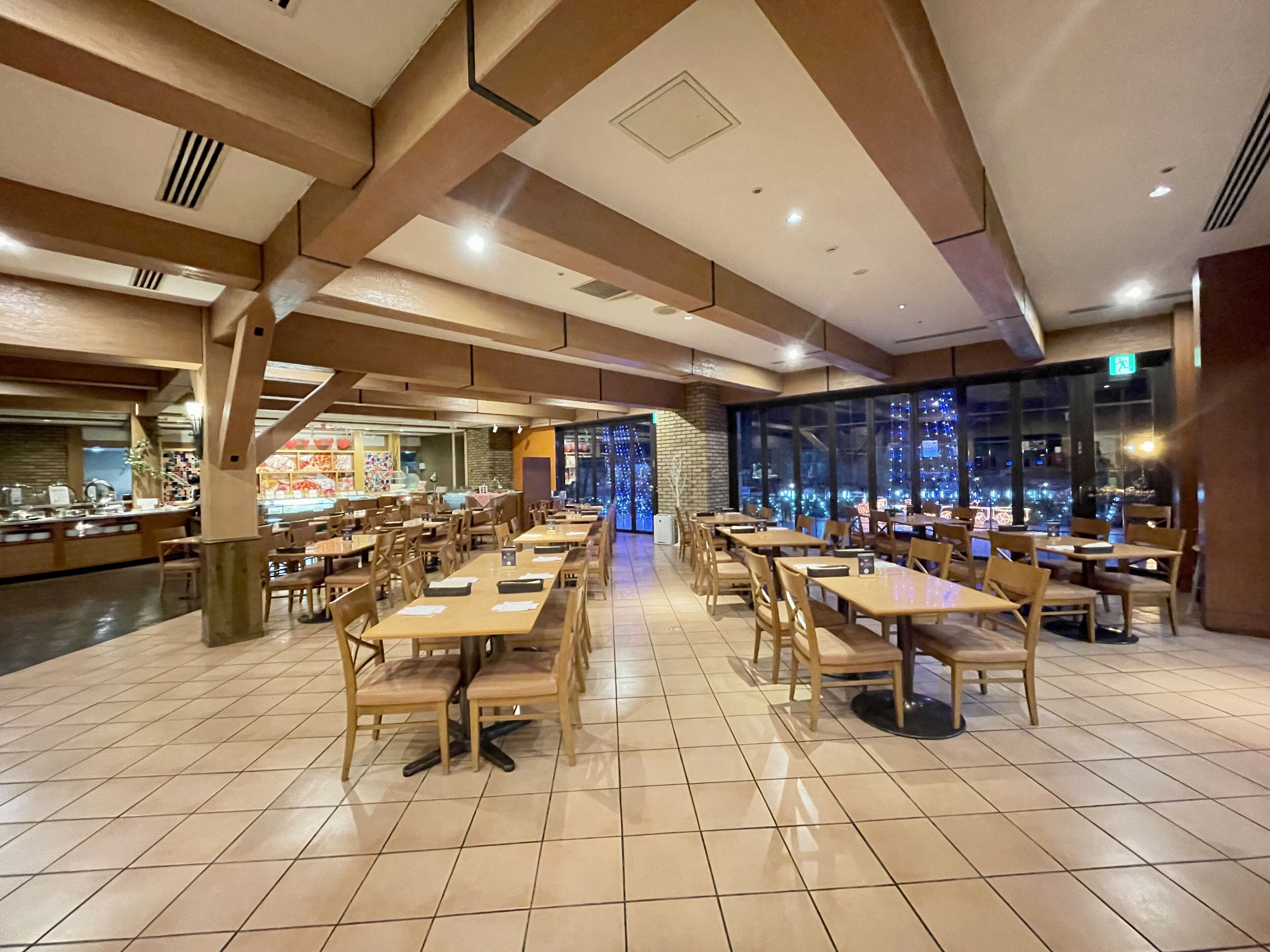 A spacious restaurant with multiple dining tables and chairs, a kitchen area, and large windows offering a city view.