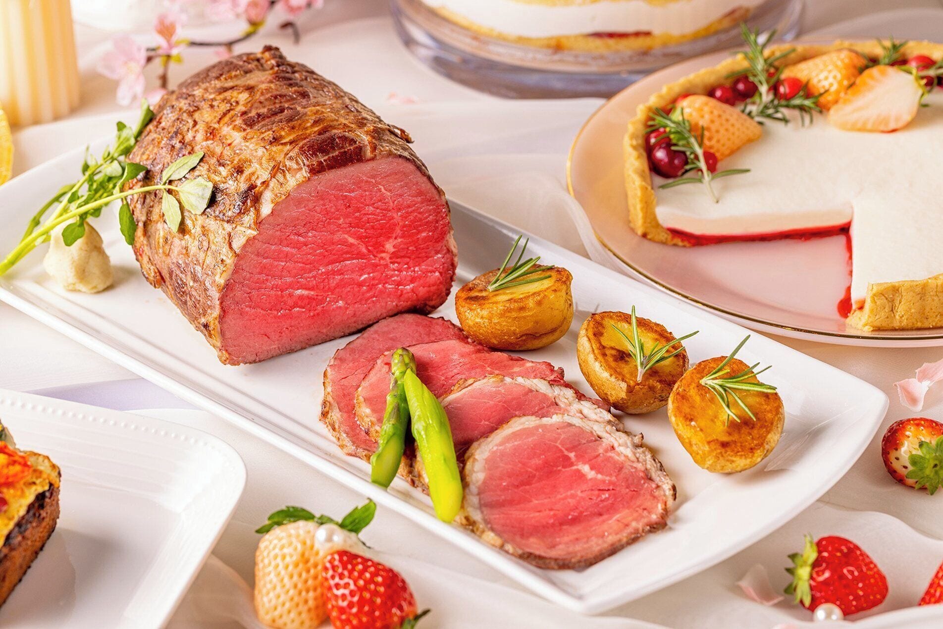 Un plato principal de carne asada acompañado de espárragos y papas al horno, junto con un postre y fresas.