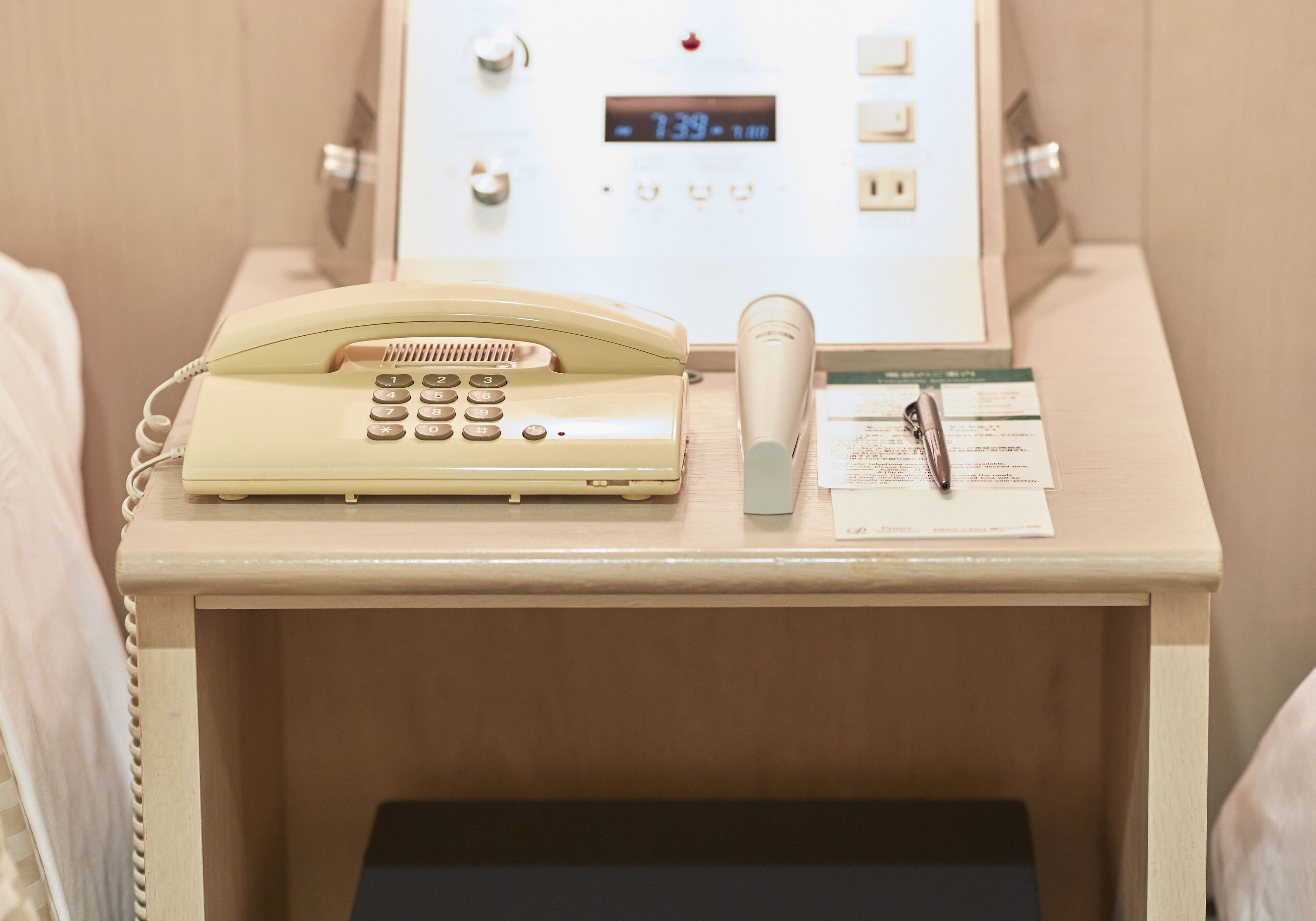 Una mesita de noche con un teléfono de estilo vintage, un reloj digital moderno y un bloc de notas con un bolígrafo.