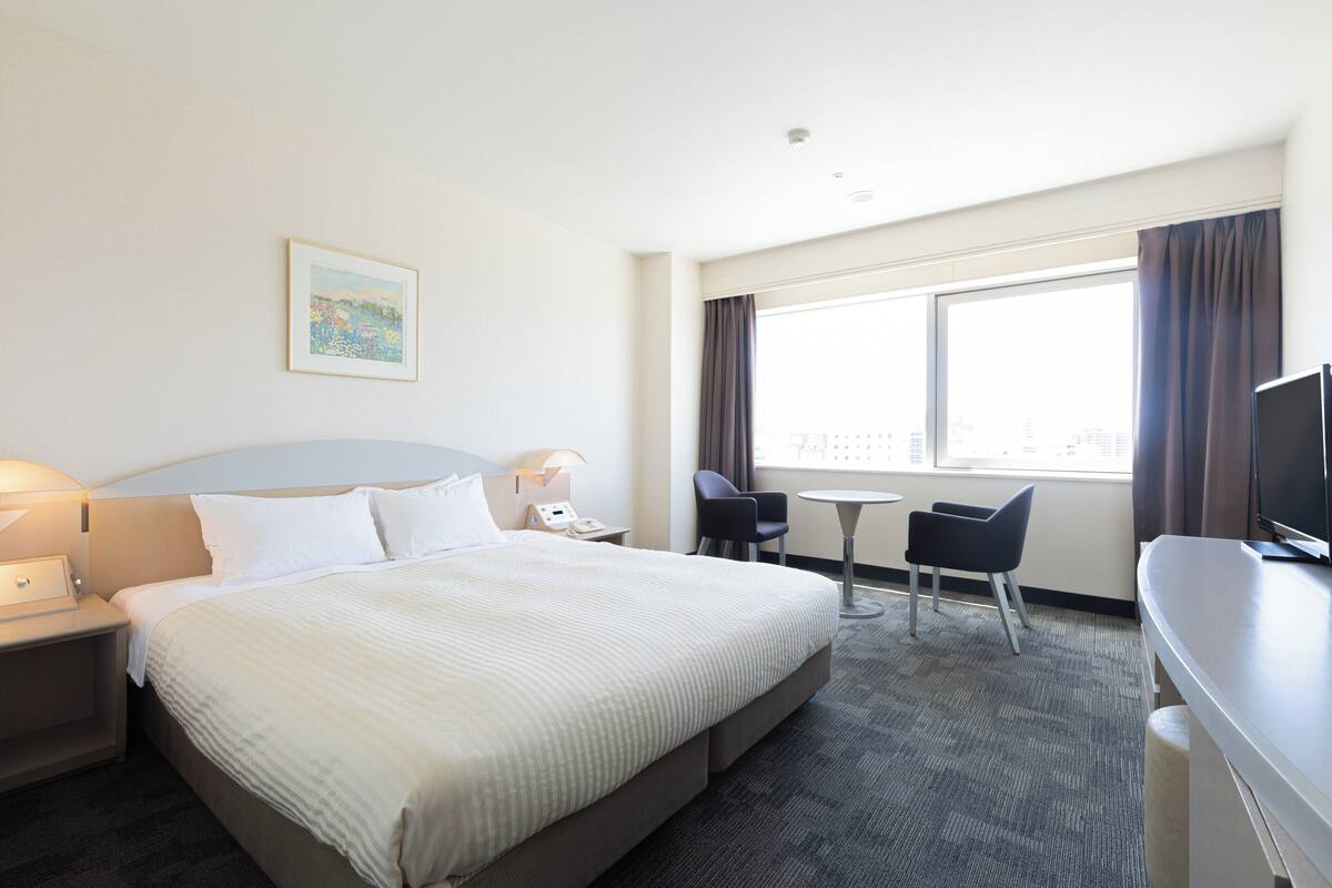 A hotel room with a large bed, a desk, a chair, a small table, and a television.