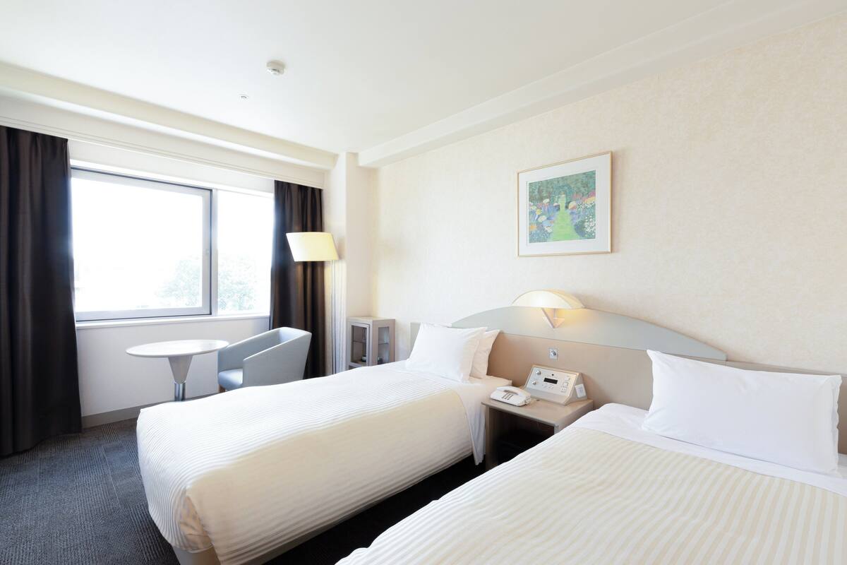 A hotel room with two beds, a desk, a chair, a lamp, and a painting on the wall.