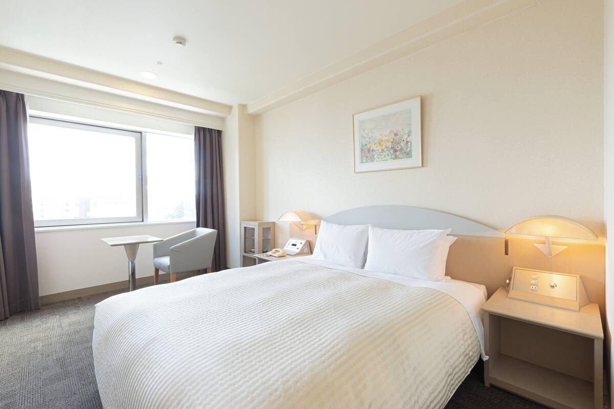 A hotel room with a large bed, a desk, a chair, a nightstand, and a wall-mounted lamp.