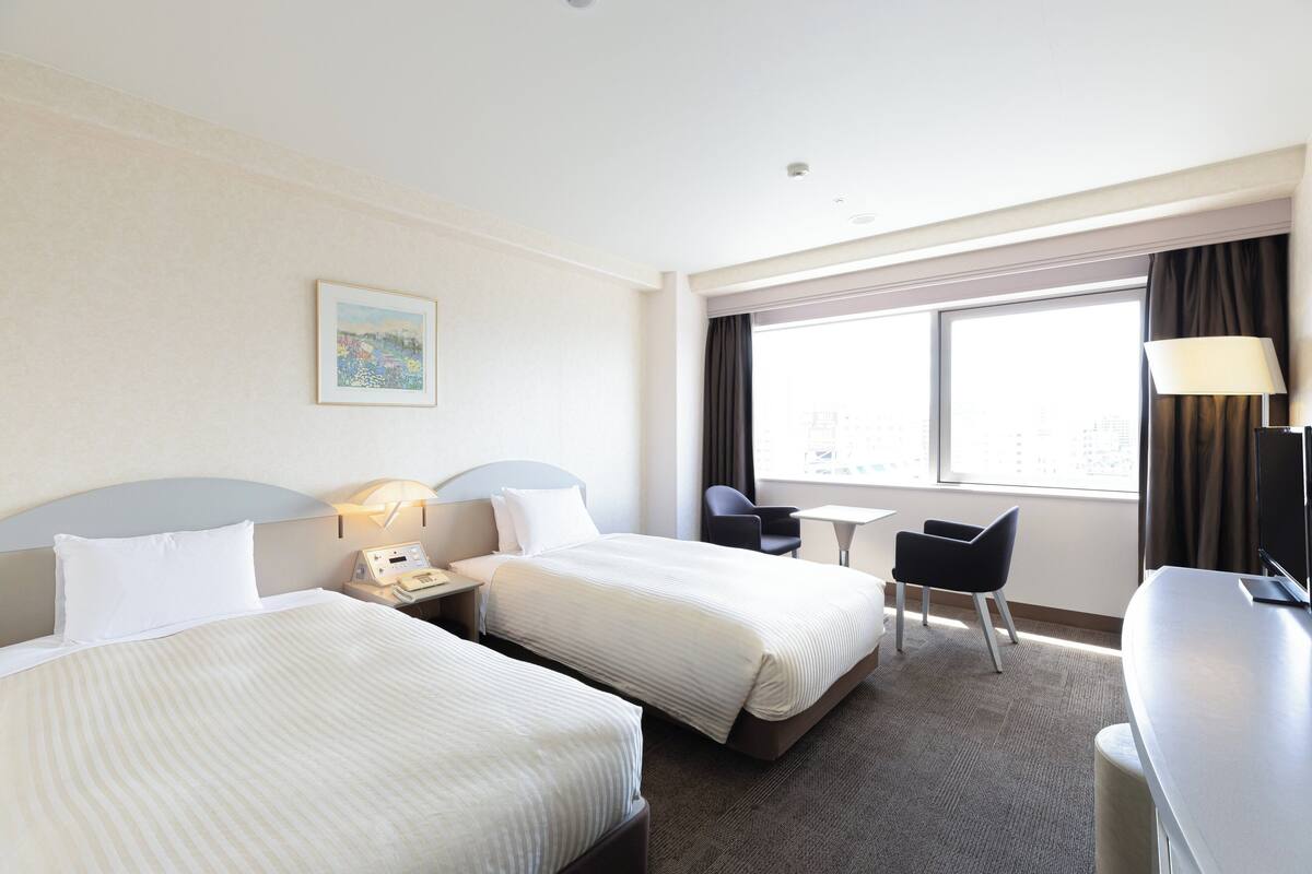 A hotel room with two beds, a desk, a chair, a small table, and a window with curtains.