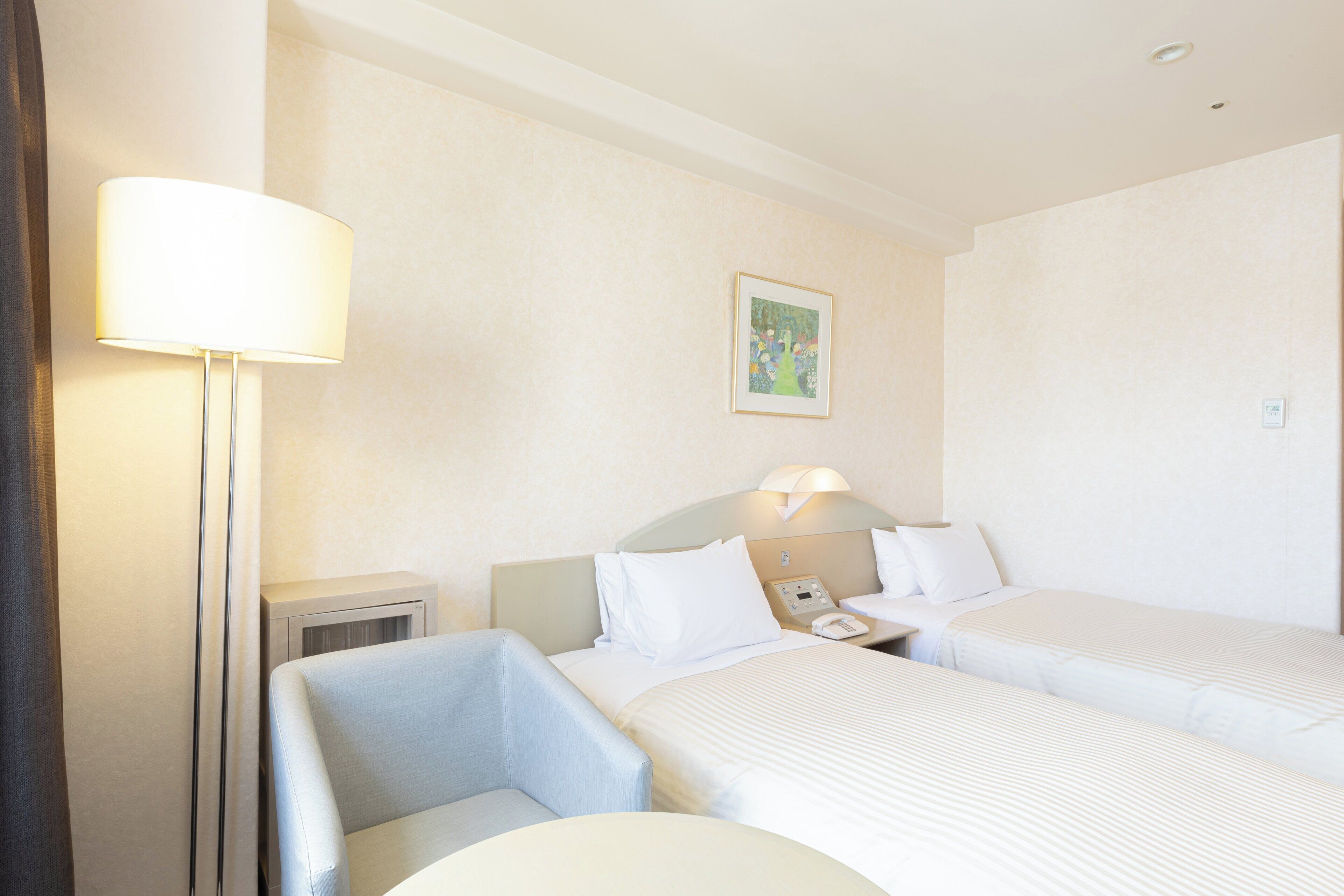 Habitación de hotel con dos camas, una mesita de noche con lámpara, una silla y un cuadro en la pared.