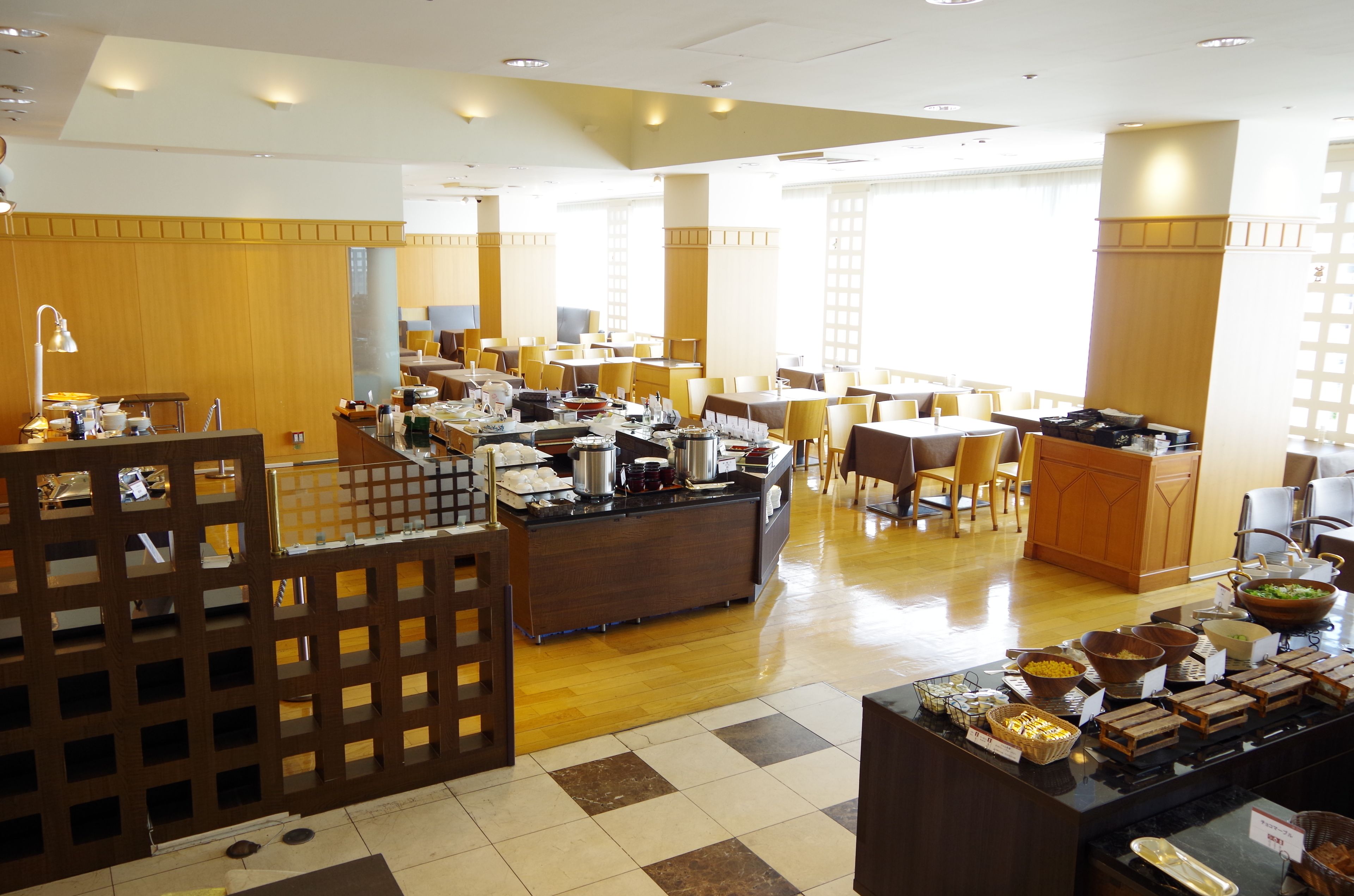 A spacious hotel dining area with multiple tables and chairs, a buffet counter with various food items, and a wooden partition with a grid design.