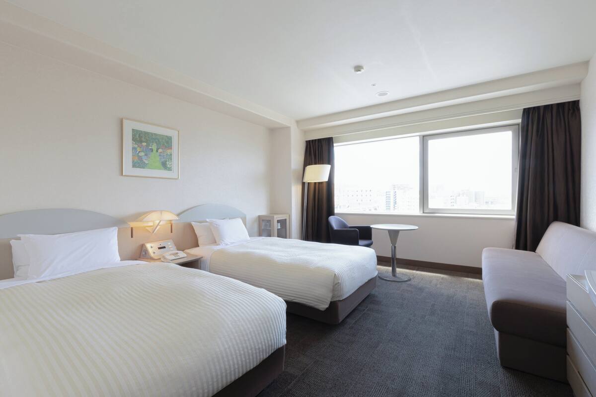 A hotel room with two beds, a desk, a chair, a sofa, a lamp, and a window with curtains.