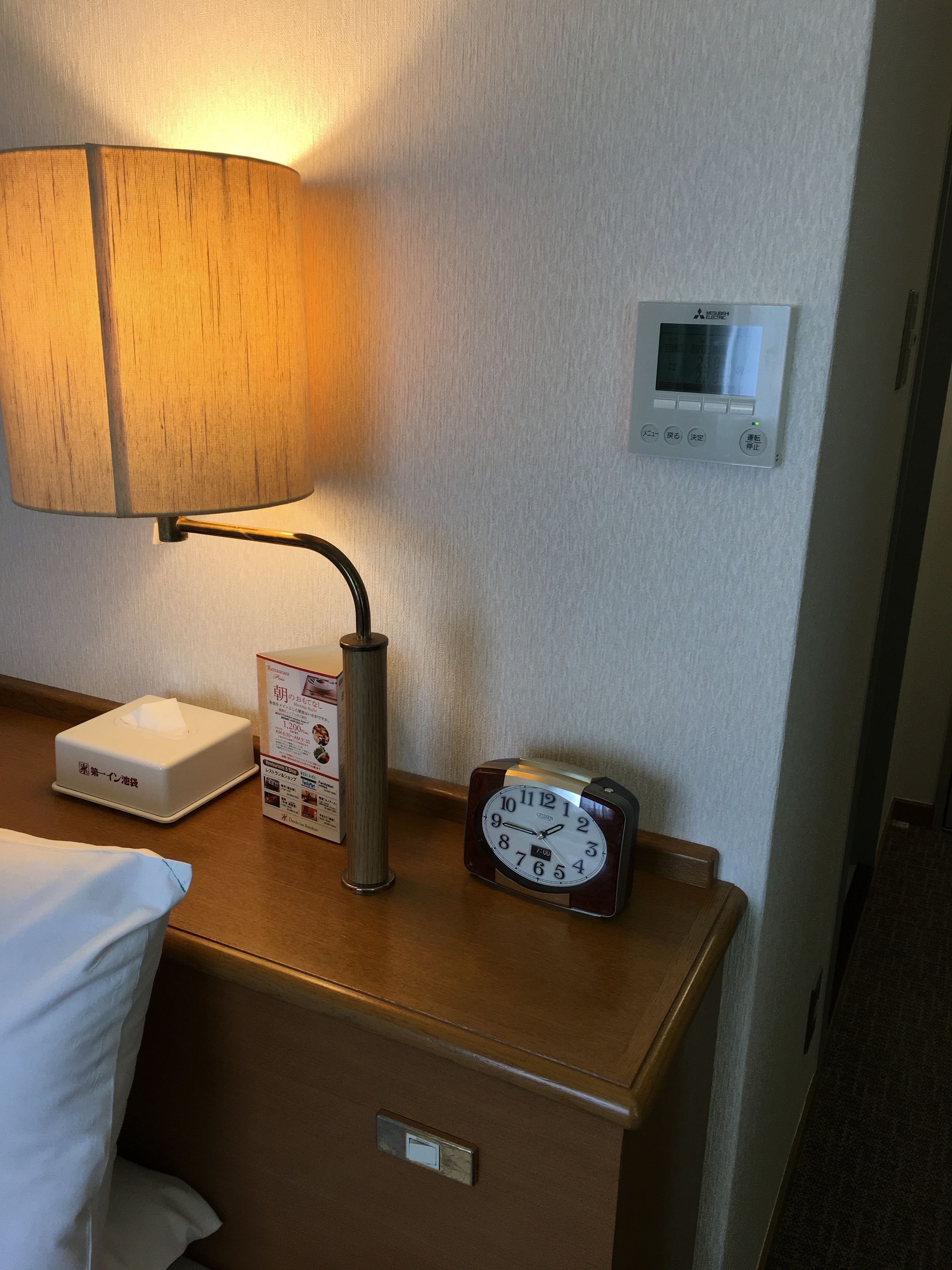 Een hotelkamer met een bedlamp, een zakdoos, een digitale thermostaat en een klassieke analoge klok op het nachtkastje.