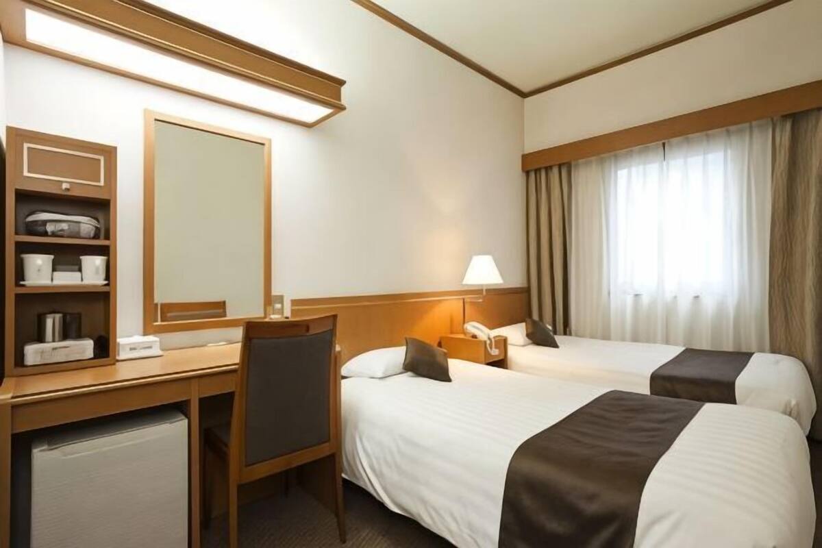 A hotel room with two beds, a desk, a chair, a mirror, a lamp, and a window with curtains.