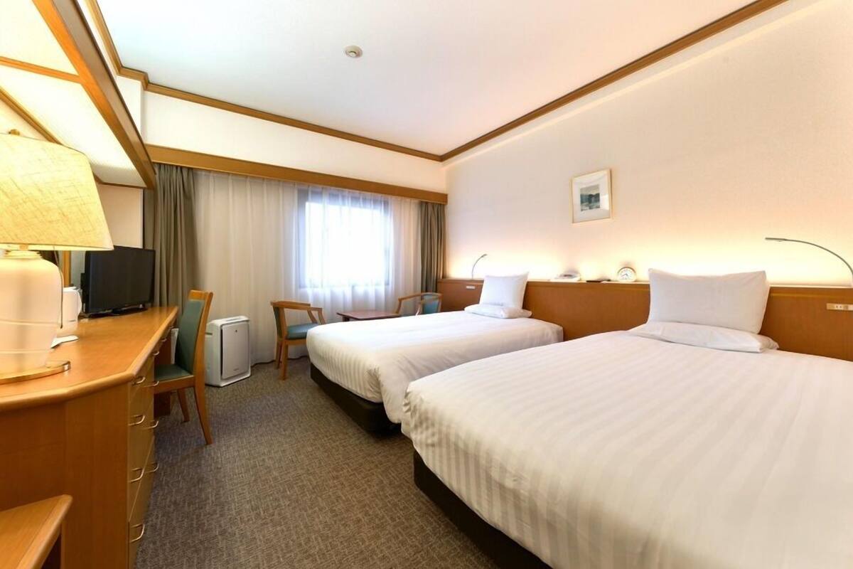 A hotel room with two beds, a desk with a lamp, a chair, a television, and a window with curtains.
