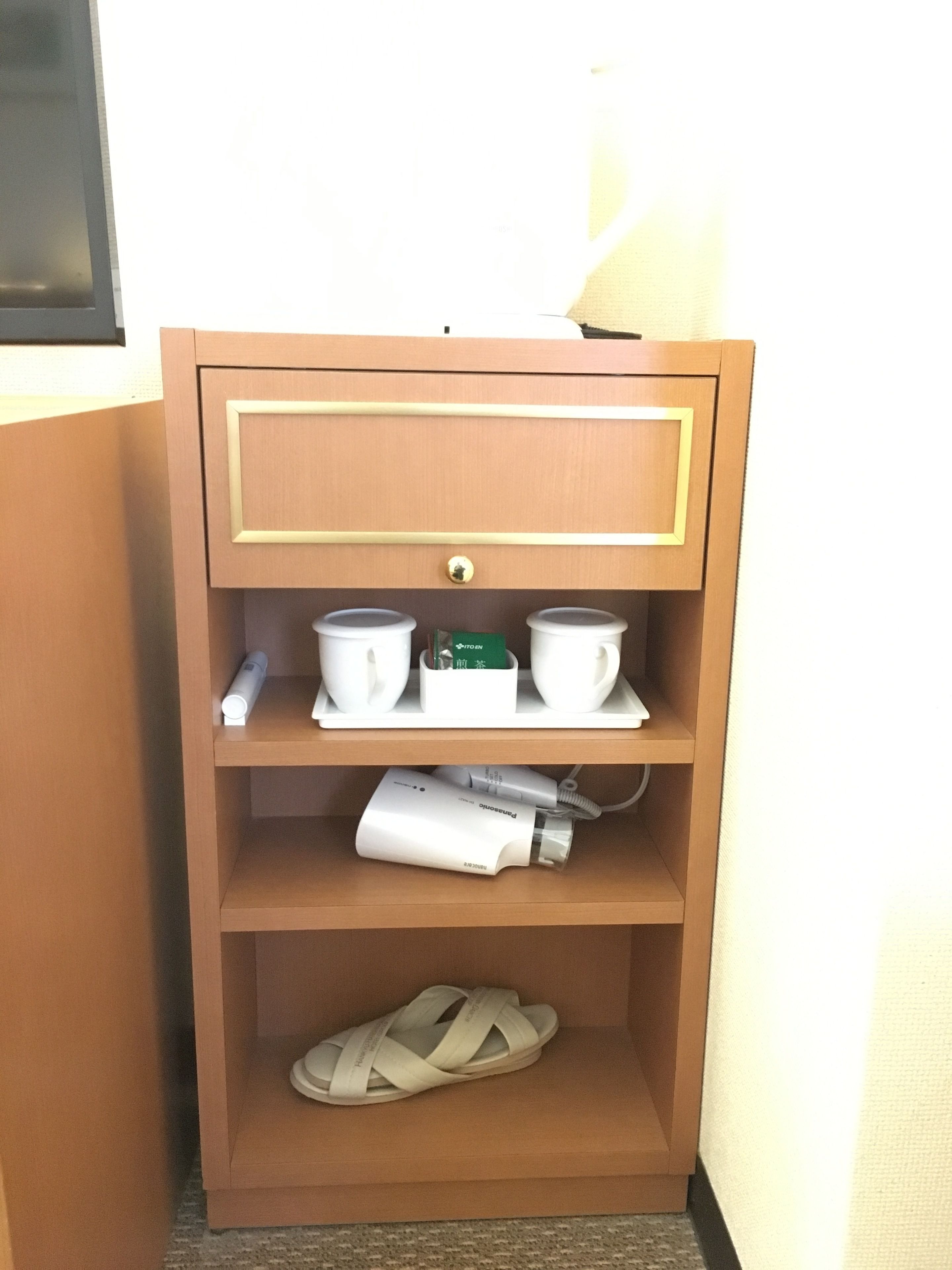 Un mueble de madera con tres estantes; el estante superior tiene dos tazas blancas y un objeto verde, el estante central tiene un dispositivo blanco y una taza blanca, y el estante inferior contiene una sandalia blanca.