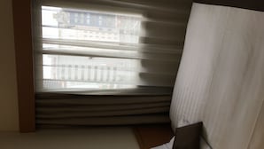 Una habitación con una cama, una ventana con persianas y vistas a edificios.