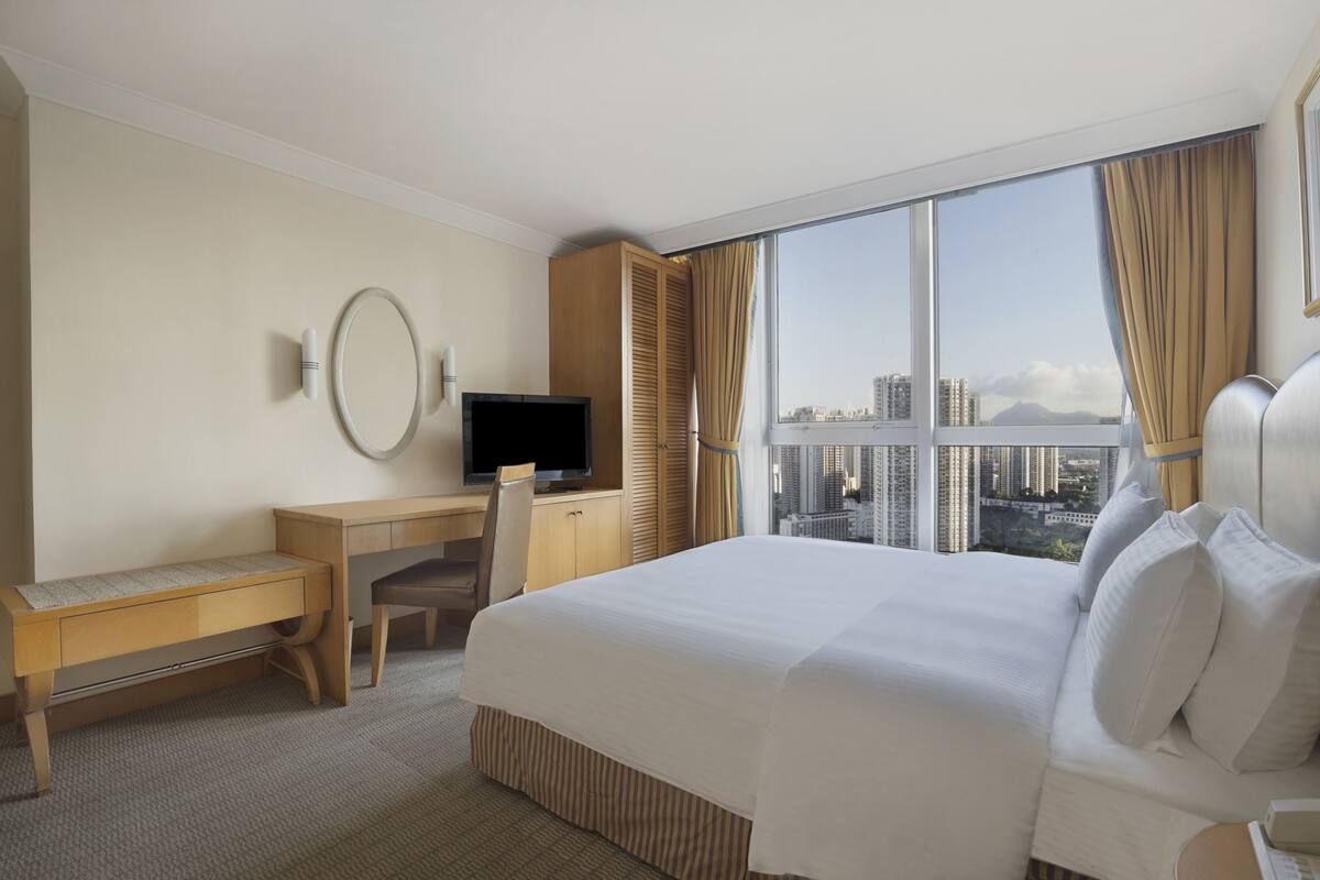Ein Hotelzimmer mit einem großen Bett, einem Schreibtisch mit Stuhl, einem Fernseher und Blick auf eine Stadtkulisse durch große Fenster.