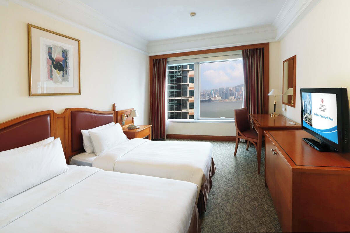 A hotel room with two beds, a desk, a television, and a large window with a city view.