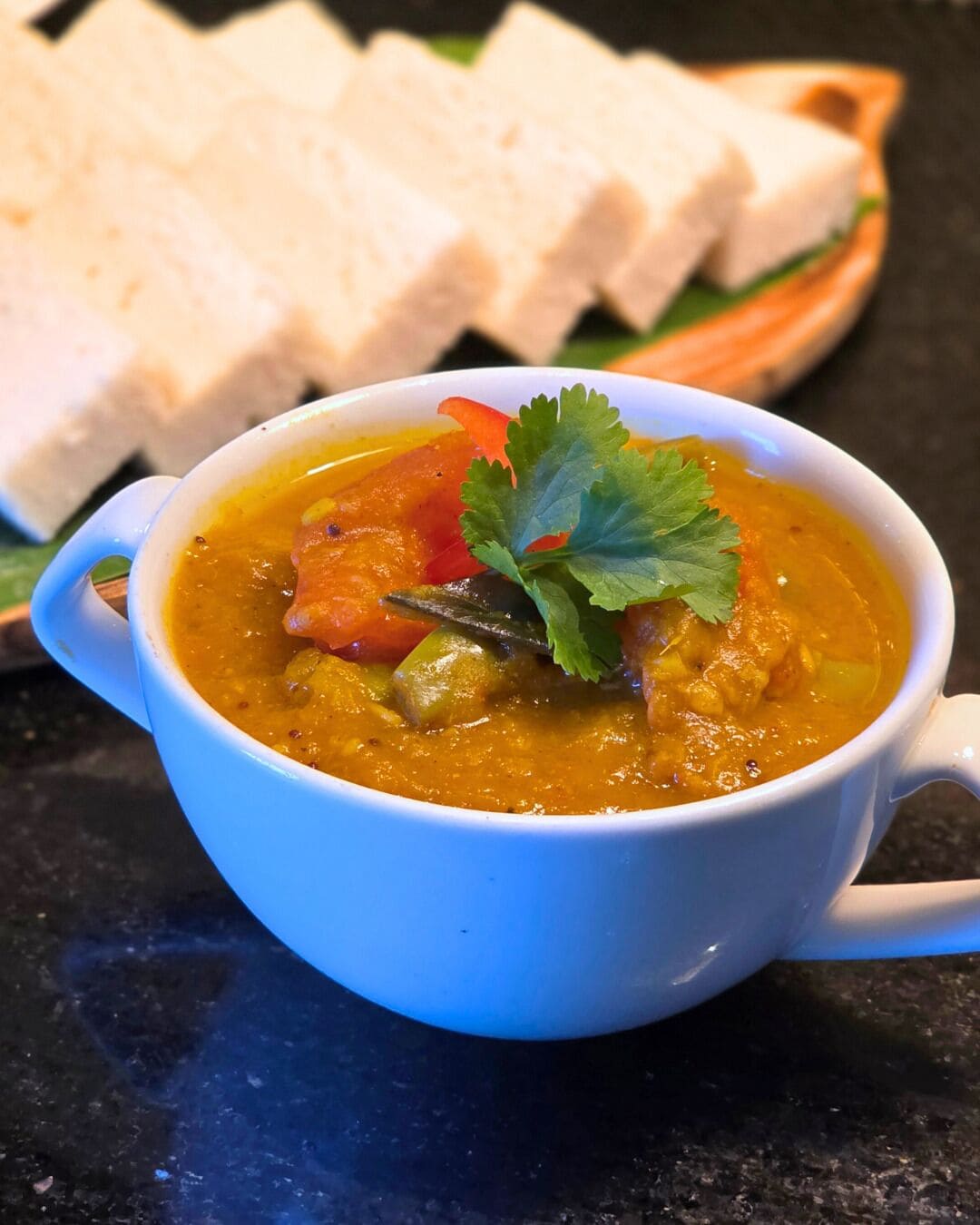 Un tazón de curry con verduras y decoración, sobre una mesa con un sándwich al fondo.