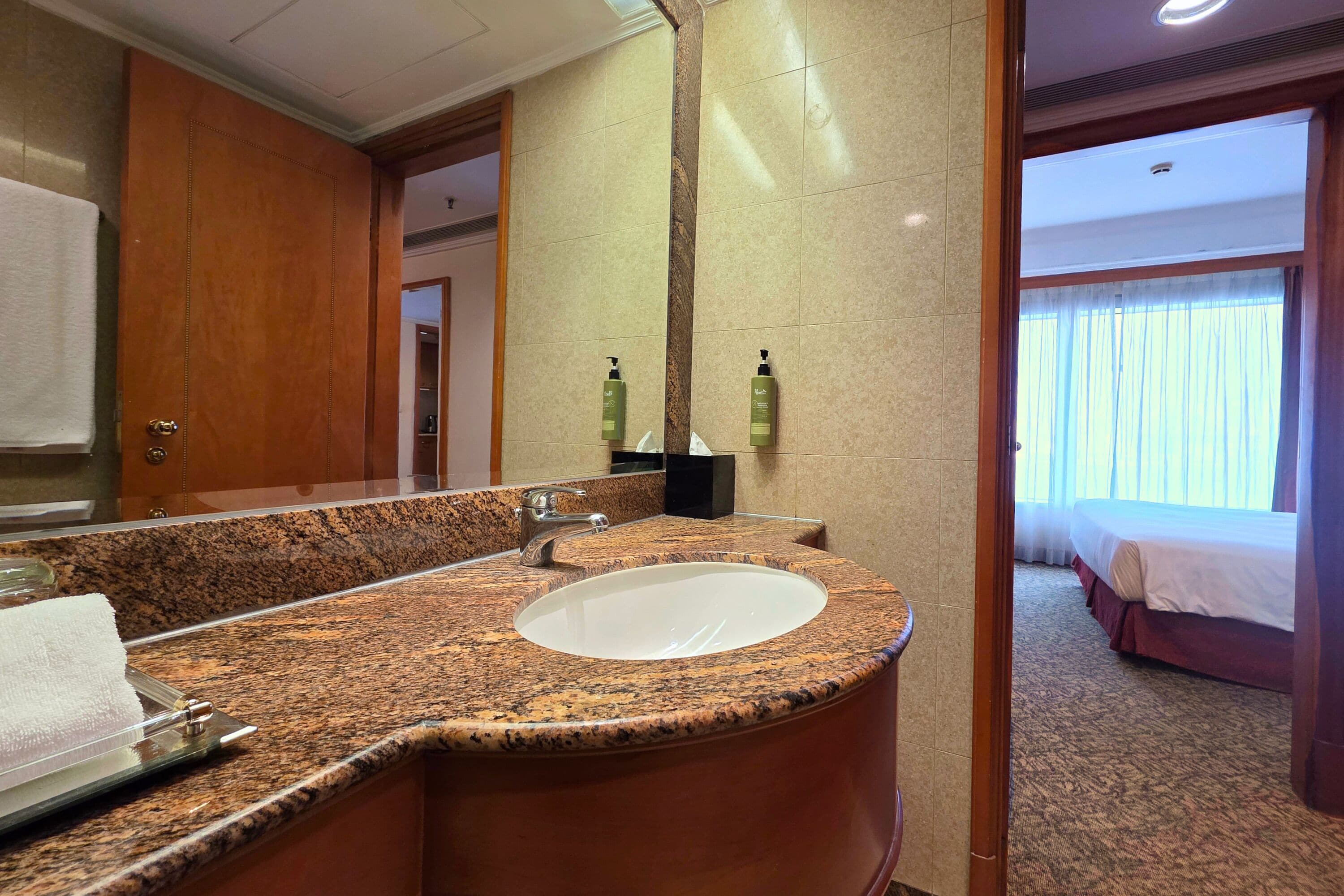 A hotel bathroom with a granite countertop sink, a mirror, and two bottles of shampoo.