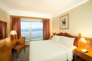 A hotel room with a large bed, a desk, a chair, and a window with a city view.