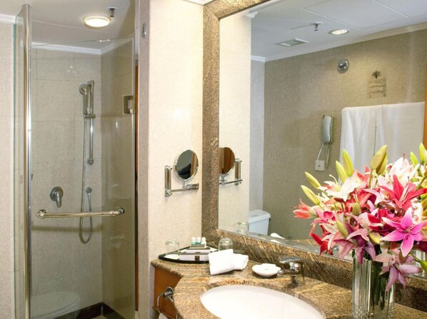 Baño de hotel con ducha de mampara de vidrio, lavamanos con encimera de granito, un espejo grande y un florero con flores.