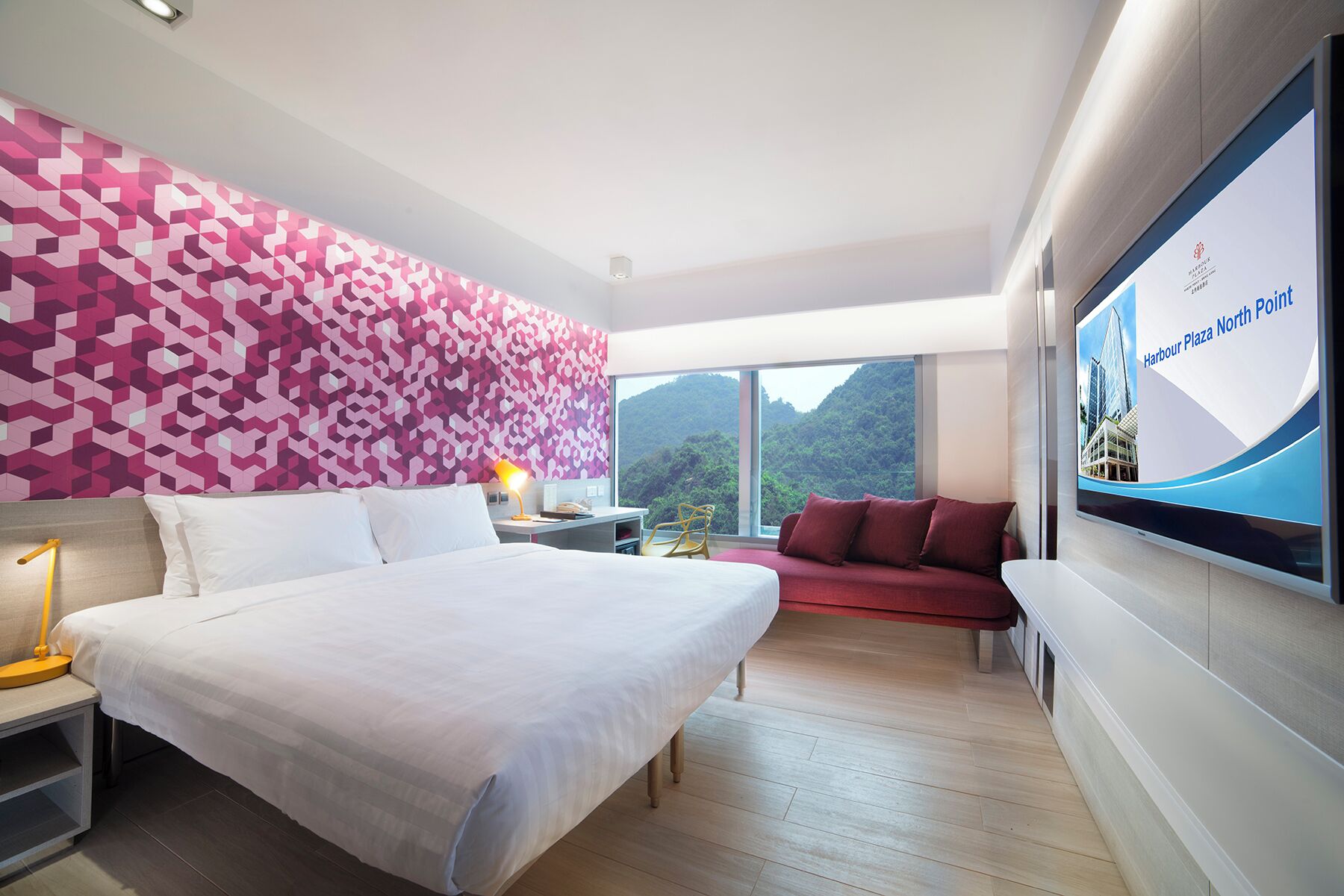 Een moderne hotelkamer met een groot bed, een rode bank, een televisiescherm waarop 'Harbour Plaza North Point' staat, en uitzicht op groene heuvels door het raam.