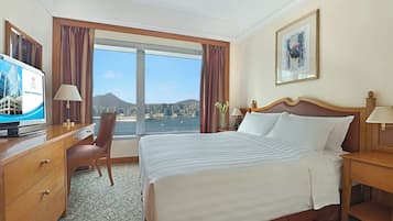 A hotel room with a large bed, a desk with a television, a chair, and a view of a cityscape through large windows.