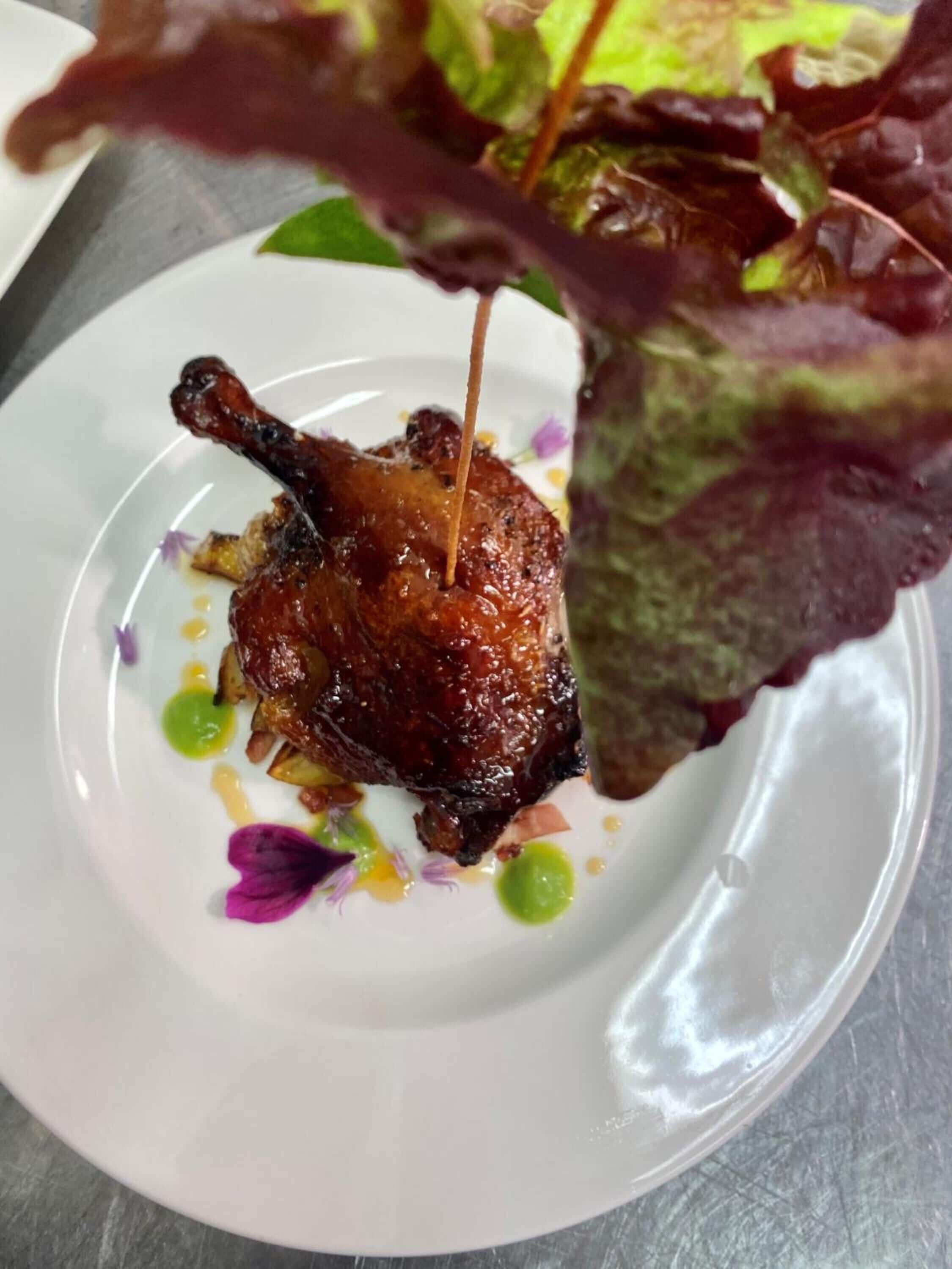 A plate with a roasted duck leg, garnished with edible flowers and a drizzle of sauce.