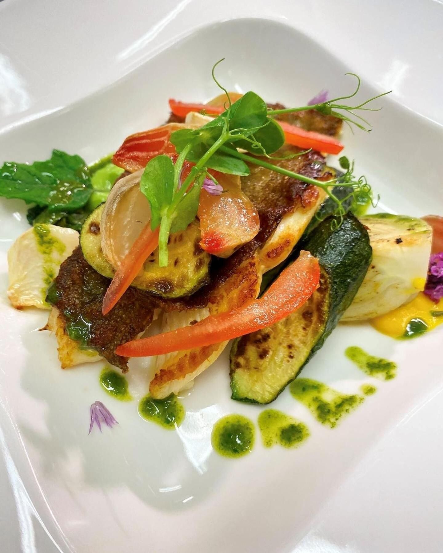 Un plato blanco con un guiso de verduras, incluyendo calabacín, pimientos rojos y hojas verdes, adornado con microbrotes y una salsa verde.