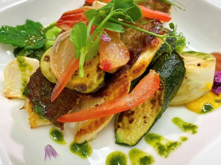 Un plato blanco con un guiso de verduras, incluyendo calabacín, pimientos rojos y hojas verdes, adornado con microbrotes y una salsa verde.
