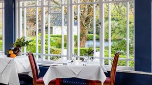 Un coin repas avec une table dressée pour deux personnes, de grandes fenêtres donnant sur un jardin et un bâtiment, et un radiateur placé sous la table.
