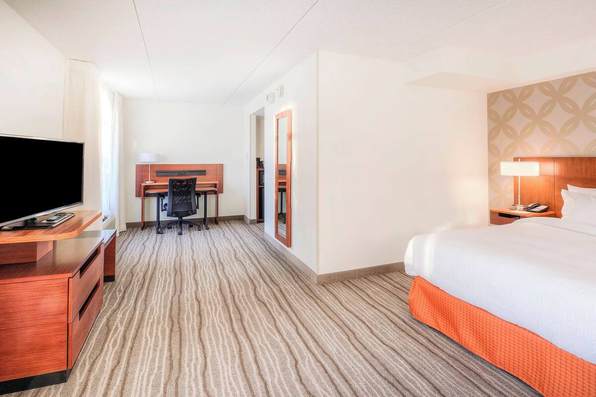 Habitación de hotel con cama, escritorio, televisor y baño visible a través de una puerta abierta.