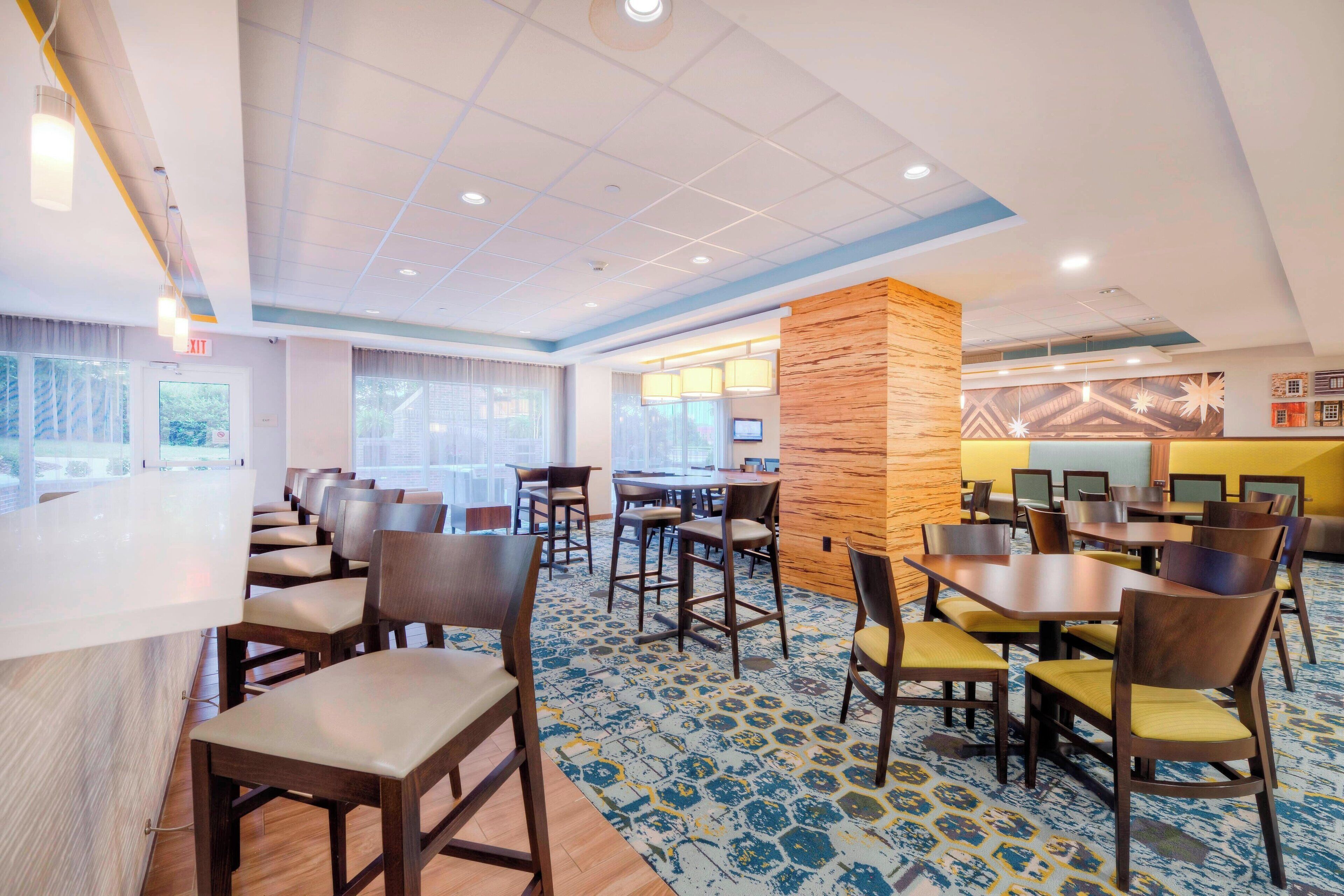 Eine Hotelhalle mit einer langen Rezeption, mehreren Sitzbereichen mit Tischen und Stühlen sowie großen Fenstern, die viel Tageslicht hereinlassen.