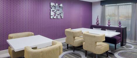 A hotel room with a purple patterned wall, a white table, beige chairs, and a purple sofa.