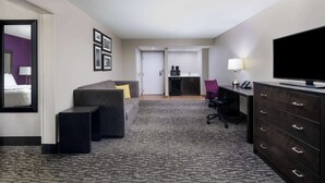 A hotel room with a sofa, a flat-screen TV, a desk with a lamp, a coffee machine, and a small kitchenette.