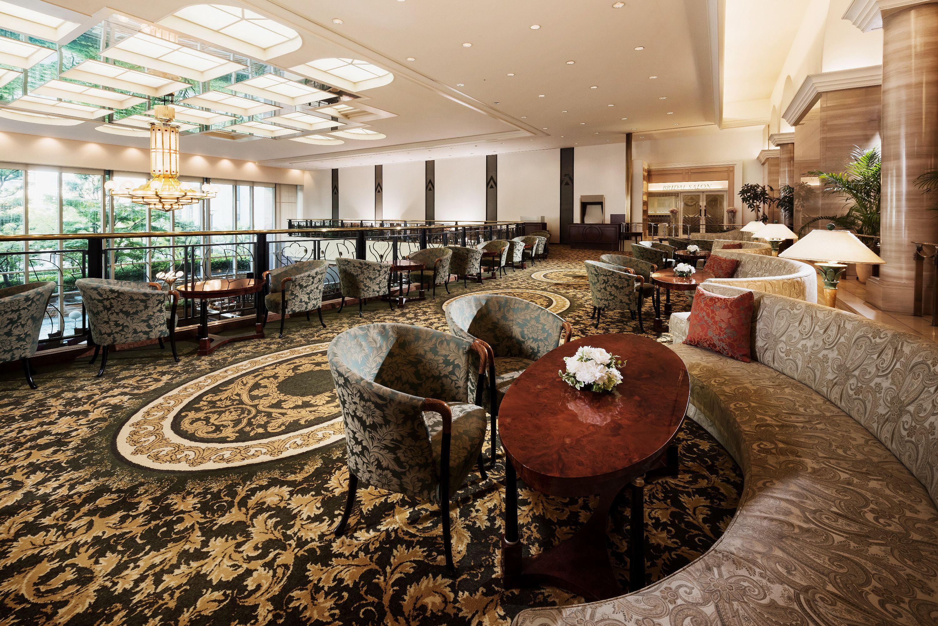 A spacious hotel lobby with a high ceiling, large windows, and a central seating area with sofas and armchairs.