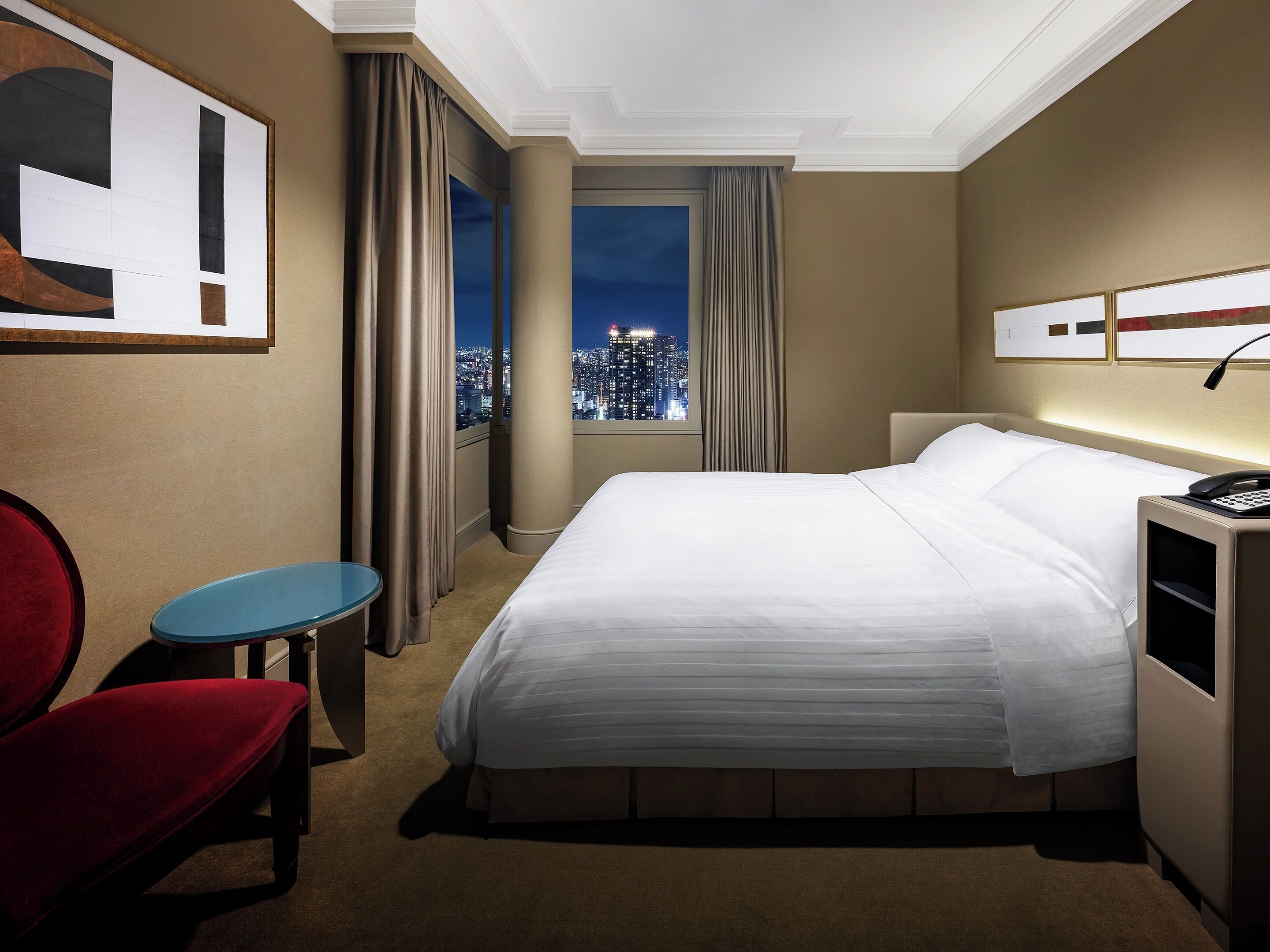 A hotel room with a large bed, a desk, a chair, and a view of a cityscape at night.