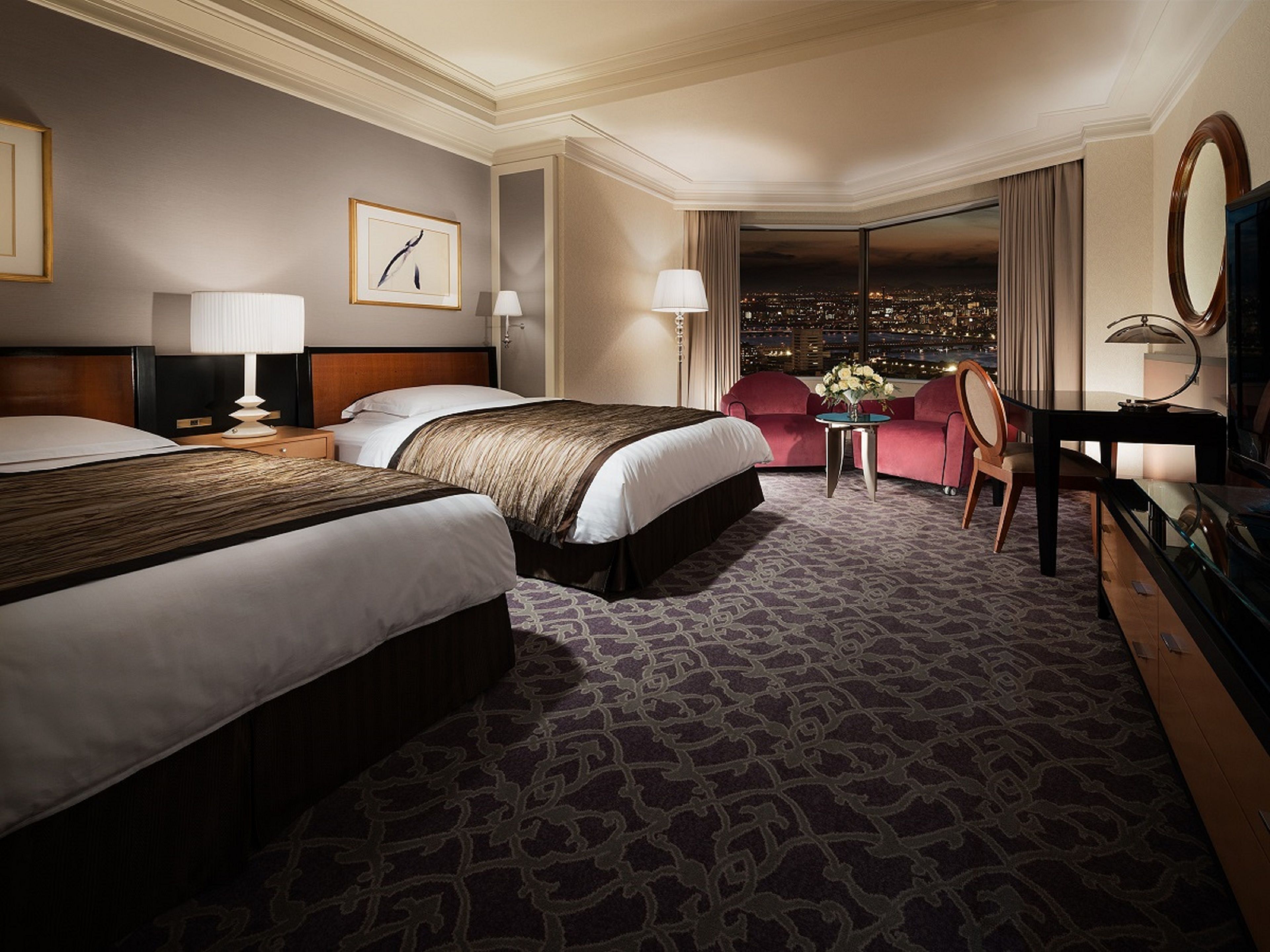 A hotel room with two beds, a desk, a chair, and a view of the city at night.