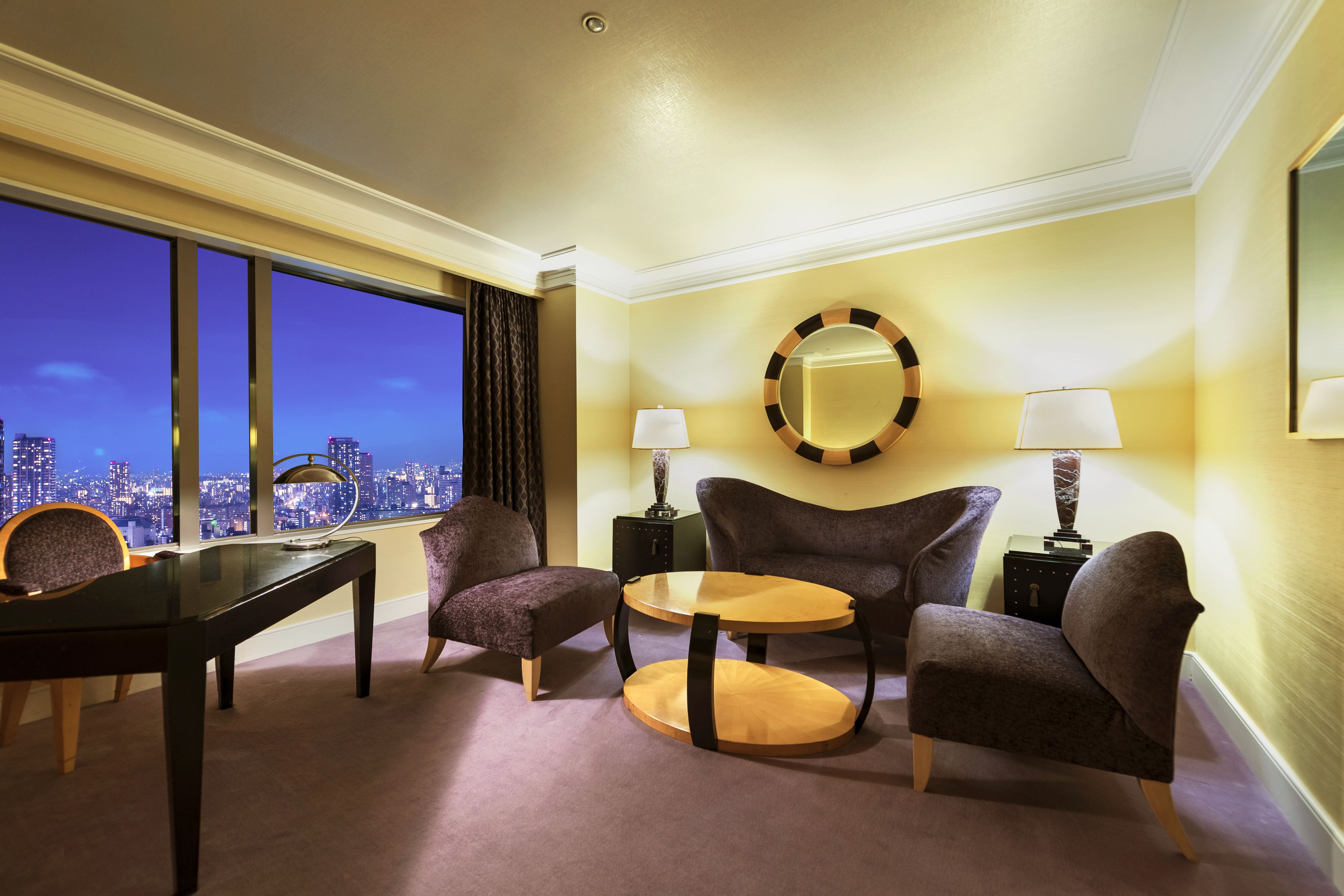 A hotel room with a city view, two armchairs, a small round table, a desk, and two lamps.