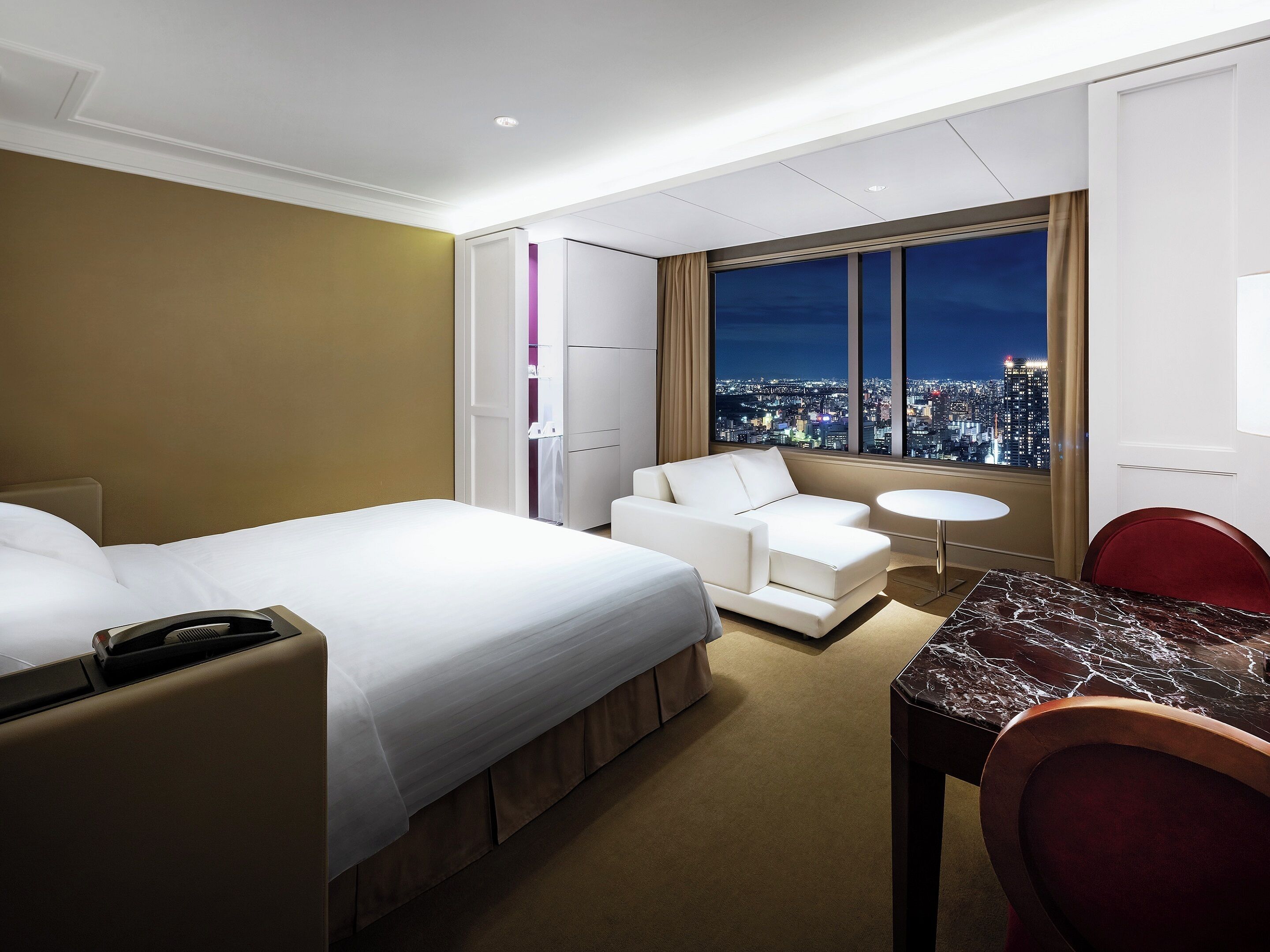 Ein Hotelzimmer mit Bett, einer Couch, einem Esstisch und Blick auf eine nächtliche Stadtansicht.